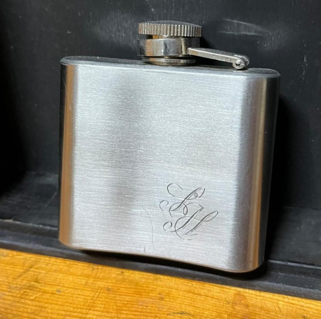 Das Bild zeigt einen Flachmann aus Stahl vor einem schwarzen Hintergrund. Verziert wird er durch ein handgraviertes Monogramm, was die sanft geschwungene und leicht ineinander verschlungenen Buchstaben SH zeigt.