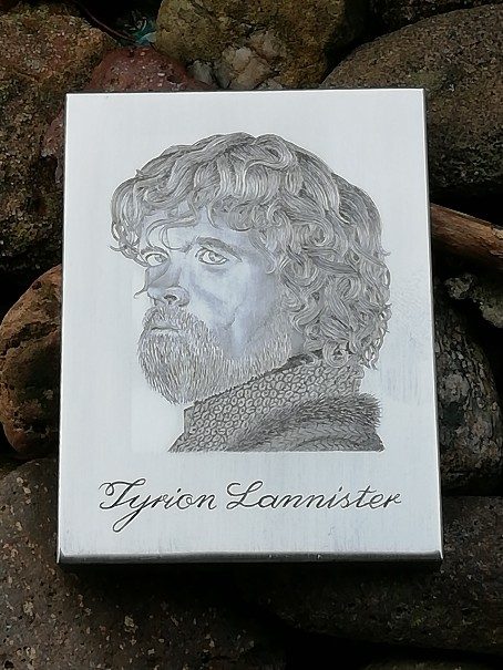 Das Bild zeigt ein graviertes Porträt von Tyrion Lannister, einer Figur aus der Serie "Game of Thrones", gespielt vom Schauspieler Peter Dinklage. Das Porträt ist detailliert ausgearbeitet und zeigt Tyrion mit seinem markanten Gesichtsausdruck, lockigem Haar und Bart. Darunter ist in geschwungener Schrift der Name "Tyrion Lannister" graviert. Der Hintergrund des Bildes besteht aus unregelmäßig geformten Steinen, auf denen die Platte mit der Gravur liegt, die dem Bild eine rustikale Atmosphäre verleihen.