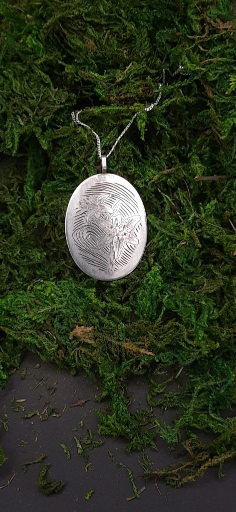 Das Bild zeigt ein ovales Medaillon mit Handgravuren, das an einer silbernen Kette hängt. Die Gravur auf dem Medaillon zeigt eine Lilie, die Blüten und Blätter sind fein gestochen, wobei die sanften Linien die Konturen und Texturen der Blumen betonen. Hinter der Blume ist ein Fingerabdruck graviert. Der Kontrast zwischen dem glänzenden Silber des Medaillons und dem natürlichen Grün des Mooses, auf dem der Anhänger liegt, hebt die handwerkliche Präzision der Gravur hervor.