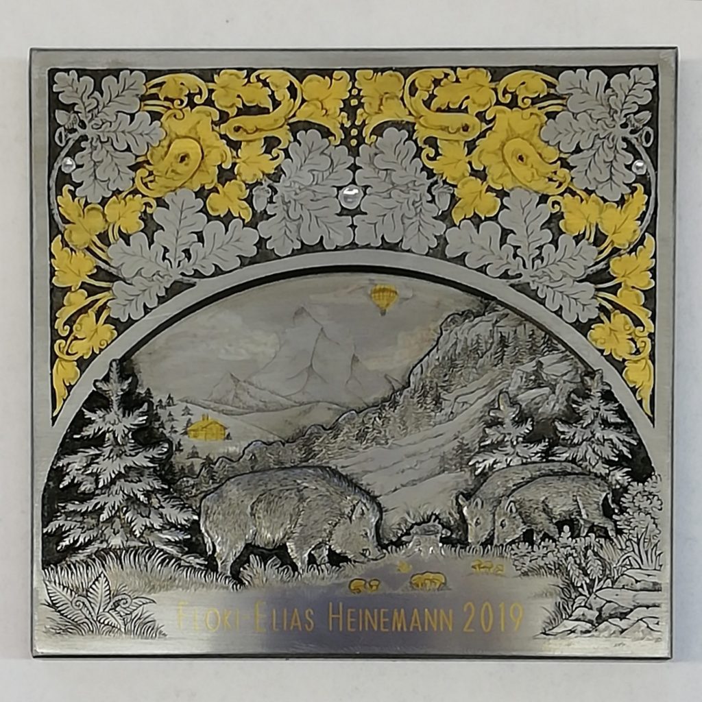 Das Bild zeigt eine kunstvolle Reliefgravur auf einer rechteckigen Metallplatte mit mehreren Ebenen. Das Relief befinde sich auf der unteren Hälfte der Platte in einem Halbkreis. Sie zeigt eine detailliert gravierte Waldszene. Im Vordergrund stehen drei Wildschweine auf einer Lichtung, auf der Pilze wachsen, die vergoldet wurden und links und rechts stehen Tannen. Der Mittelgrund zeigt einen Hang, hinter dem ein Wald in der Ferne steht, während im Hintergrund Berge zu sehen sind, mit einer kleinen vergoldeten Holzhütte. Über der Szene fliegt ein ebenfalls vergoldeter Heißluftballons am Himmel. Die obere Hälfte der Platte ist verziert mit zwei miteinander kombinierte Ornament, welche in der Mitte vertikal symmetrisch sind. Die verwendeten Ornamente sind Wetzlaub, welches vergoldet wurde und Eichenlaub. Am unteren Rand der Platte ist in Gold tauschierter Schrift der Name "Floki-Elias Heinemann 2019".