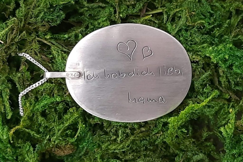 Das Bild zeigt ein ovales Medaillon mit von Hand gravierter Schrift, welches an einer silbernen Kette hängt. Der Schriftzug liest: „Ich hab dich lieb, Mama“ und ist nach einer Schriftvorlage der Handschrift der Mutter graviert. Oben drüber sind zwei von Handgezeichnete Herzen graviert. Der Kontrast zwischen dem glänzenden Silber des Medaillons und dem natürlichen Grün des Mooses, auf dem der Anhänger liegt, hebt die handwerkliche Präzision der Gravur hervor.