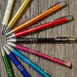 Kugelschreiber in verschiedenen Farben liegen in einem Halbkreis angeordnet. Der mittlere Kugelschreiber ist versehen mit einer Handgravur. Die Gravur zeigt in feiner Schreibschrift das Wort Silberstichel.