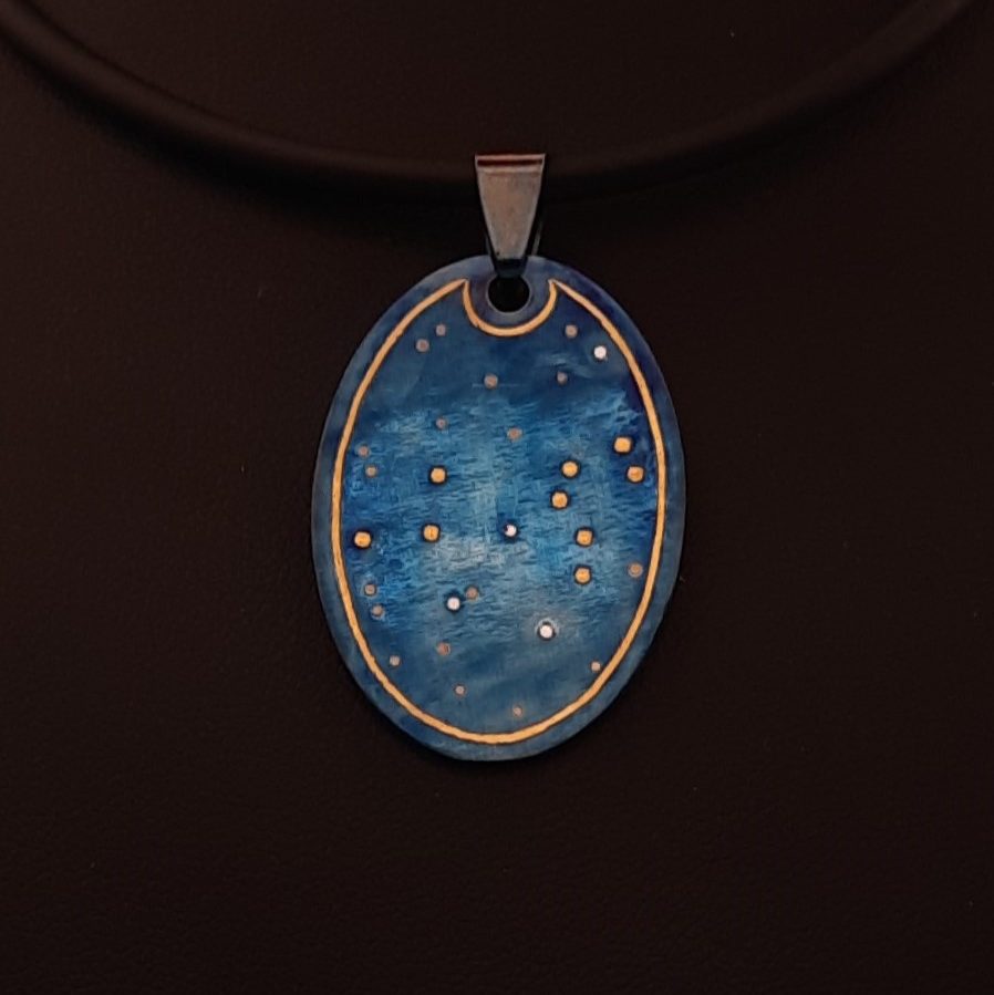 Das Bild zeigt eine Halskette mit einem ovalen Anhänger an einer schwarzen Kordel, die auf einer schwarzen Schmuckbüste präsentiert wird. Der Anhänger hat eine leuchtend blaue Oberfläche mit kleinen, silbernen und goldenen Punkten, die an Sterne am Nachthimmel erinnern. Das innere des Anhängers ist mit einer goldenen Linie umrandet.
