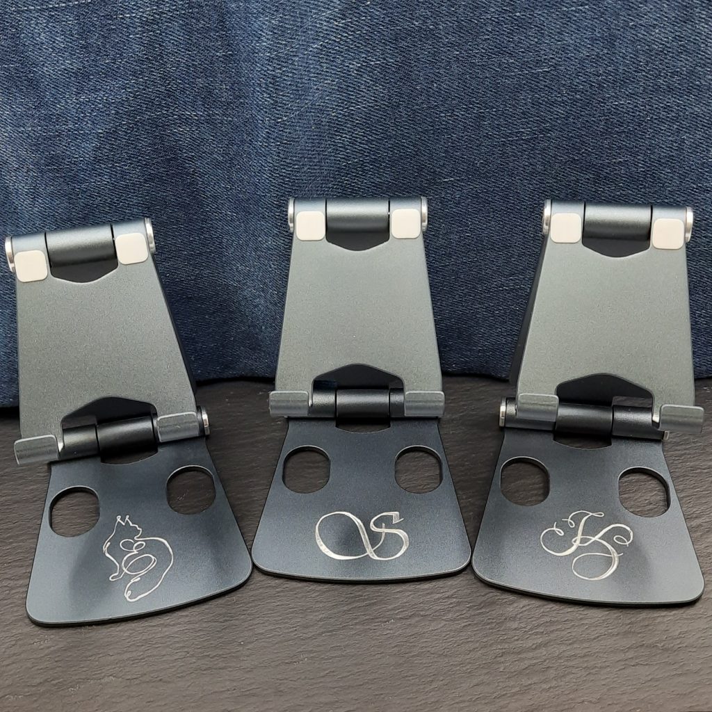 Auf dem Bild sind drei dunkelgraue, klappbare Handyständer zu sehen, die jeweils eine individuelle Gravur auf der unteren Fläche tragen. Der Hintergrund besteht aus einem dunkelblauen Jeansstoff, der den schlichten und professionellen Eindruck der Ständer unterstreicht. Linker Ständer: Dieser zeigt eine Gravur eines stilisierten Katzenmotivs, bei dem die Katze in einer geschwungenen Linie dargestellt ist, die in ein geschwungenes Sachreibschrift E übergeht. Mittlerer Ständer: Auf diesem ist ein elegantes Monogramm zu sehen, das den Buchstaben S zeigt. Es ist schlicht und symmetrisch gehalten. Rechter Ständer: Hier befindet sich die Gravur des Buchstabens "K" die in einer geschwungenen und dekorativen Schreibschrift gestaltet ist. Die Gravuren wirken präzise und fein. Die symmetrische Anordnung der Ständer auf einer dunklen, ebenen Schieferplatte trägt zu einem aufgeräumten und ansprechenden Gesamtbild bei.