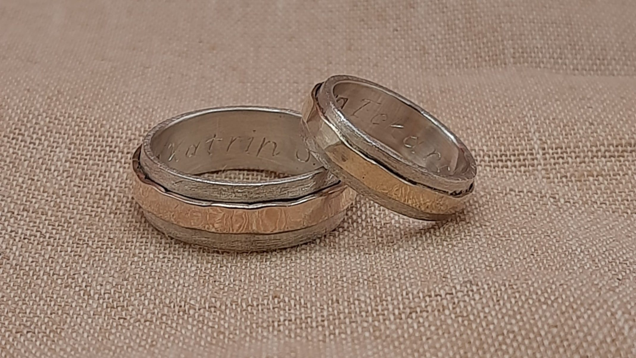 Das Bild zeigt zwei Eheringe, die auf einem beigen Stoffhintergrund liegen. Die Ringe bestehen aus einem polierten Goldring mit Hammerschlag Textur der mittig von einem breiteren Silberring mit einer gefrosteten Oberfläche erfasst wird. Der Herrenring liegt flach, während der Damenring leicht dagegen gelehnt ist, sodass die Innenseite beider Ringe sichtbar ist. In den Innenflächen der Ringe sind die Schriftzüge Eckard und Katrin eingraviert. Der Stoff im Hintergrund hat eine leicht grobe, gewebte Textur.