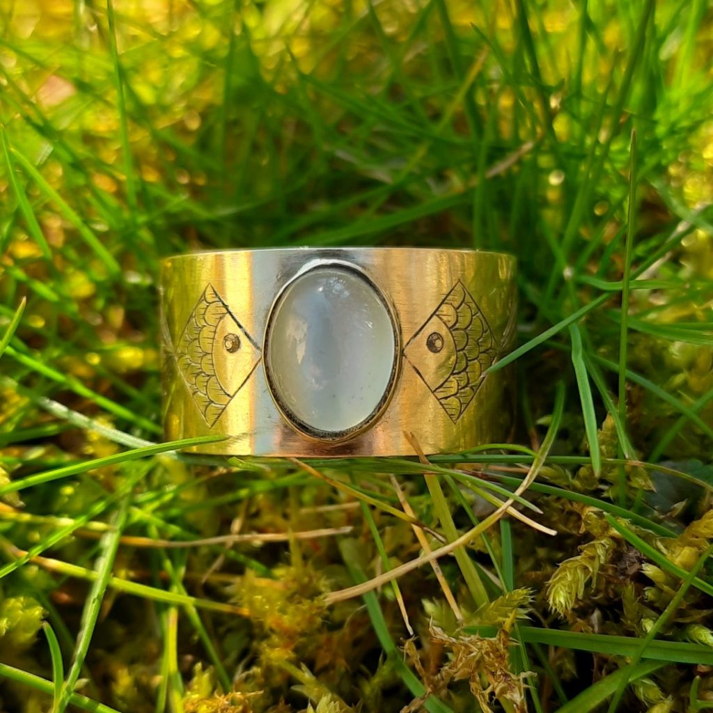 Ein Goldring mit einem ovalen Stein liegt im Gras.
Das Bild zeigt einen goldenen Ring mit einem ovalen, milchig-weißen Mondstein, der in der Mitte des Ringes eingefasst ist.
Der Ring ist breit und links und rechts vom Mondstein wurden symmetrisch vereinfachte Fische graviert. Der Ring ist so gedreht, dass man die Köpfe der Fische sehen kann. Die Fische bestehen aus einer Raute für den Körper und einem Dreieck für die Schwanzflosse. Der Körper ist mit einem Fischschuppenmuster verziert und die Schwanzflossen wurden mit strahlen artige Linien im Inneren akzentuiert. Der Fisch hat ein schwarzes rundes Auge mit einem kleinen Lichtpunkt.
Der Ring liegt auf einem Untergrund aus grünem Gras und Moos. Das Licht lässt das Grün des Grases und das Gold des Rings leuchten. Der Fokus liegt stark auf dem Ring, während der Hintergrund leicht unscharf ist und die Aufmerksamkeit auf das Schmuckstück lenkt. Die Lichtverhältnisse deuten auf einen sonnigen Tag hin.
