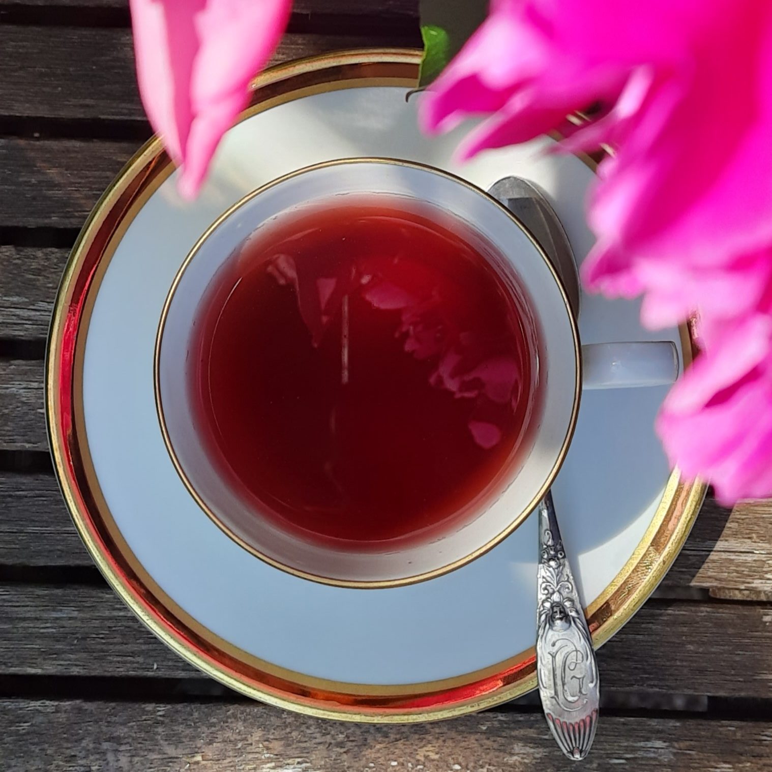 Das Bild zeigt eine elegante Teetasse von oben, die auf einer passenden Untertasse steht. Die Tasse ist mit einem tiefroten Tee gefüllt. Das Porzellan ist weiß mit einem feinen goldenen Rand und liegt auf einem rustikalen Holztisch mit sichtbarer Maserung. Ein silberner Teelöffel mit verziertem Griff liegt auf der Untertasse, rechts neben der Tasse.
Der Griff des Löffels ist reich verziert und trägt ein graviertes Monogramm in einer kunstvollen Jugendstil-Schrift – geschwungen, filigran und dekorativ. 

Von oben ragen kräftig pinkfarbene Blüten ins Bild, die den oberen Rand teilweise verdecken und eine lebendige, sommerliche Atmosphäre schaffen. Die Reflexion der Blüten ist auch auf der Oberfläche des Tees zu sehen. Das Gesamtbild wirkt ruhig, einladend und detailreich – ein Moment der Entspannung in einem blühenden Garten.